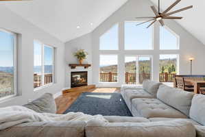 Spacious living area featuring high vaulted ceilings, expansive window walls, a built-in fireplace with a wood mantle, wood-finish flooring, and direct access to an outdoor deck