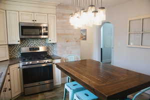 Kitchen with stainless steel appliances, cream cabinetry, backsplash, hanging lights, and arched walkways