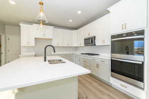 Kitchen featuring stainless steel appliances, a center island with sink, light stone counters, light wood-style flooring, and pendant lighting