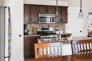 Kitchen with dark wood finish cabinets, stainless steel appliances, decorative light fixtures, tasteful backsplash, and an island with sink