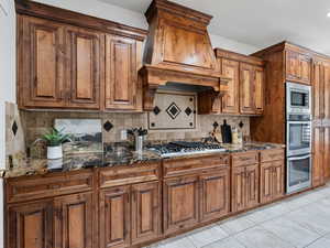 Kitchen with wood finish cabinets, dark stone counters, stainless steel appliances, tasteful backsplash, and light tile patterned floors
