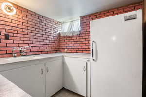 Kitchen with freestanding refrigerator, brick wall, white cabinets, and light stone counters