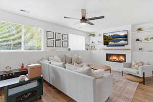 Living area with a lit fireplace, ceiling fan, light wood-type flooring, and built in features