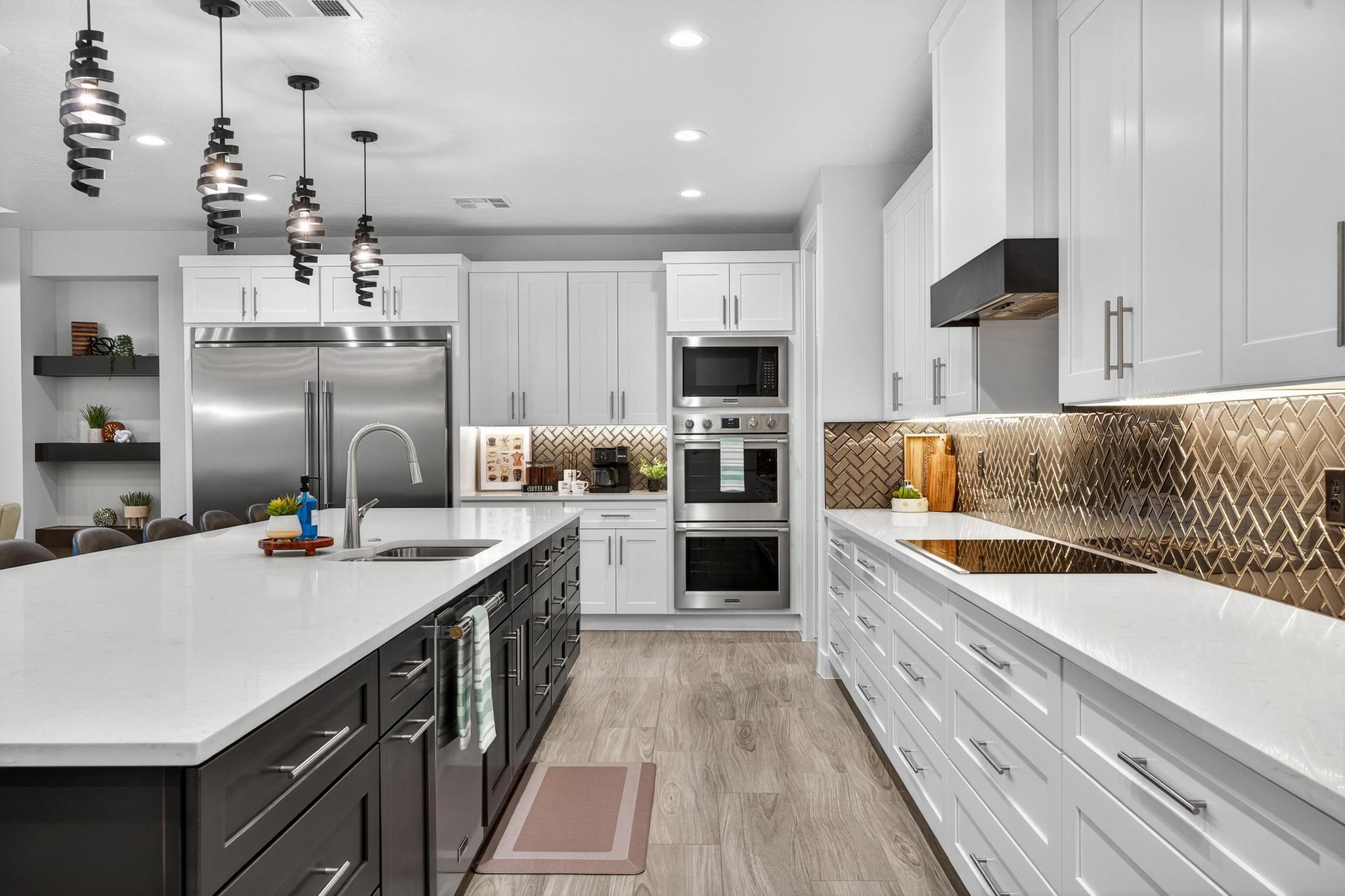 Dual tone kitchen with light stone counters, built in appliances, two tone cabinetry, decorative light fixtures, and a center island with sink