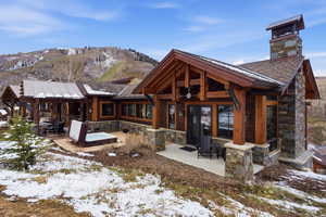 Snow covered back of property with stone siding, a patio, a mountain view, and a chimney