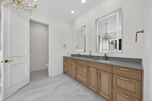 Primary suite bathroom with chandelier, double vanity, light marble finish flooring, and crown molding