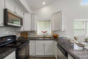 Kitchen featuring stainless steel appliances, lofted ceiling, white cabinetry, dark stone countertops, and recessed lighting