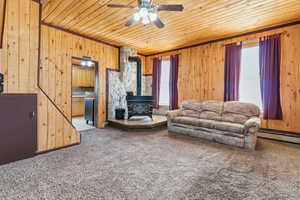 Living area featuring a wood stove, carpet flooring, a ceiling fan, wooden walls, and a baseboard heating unit