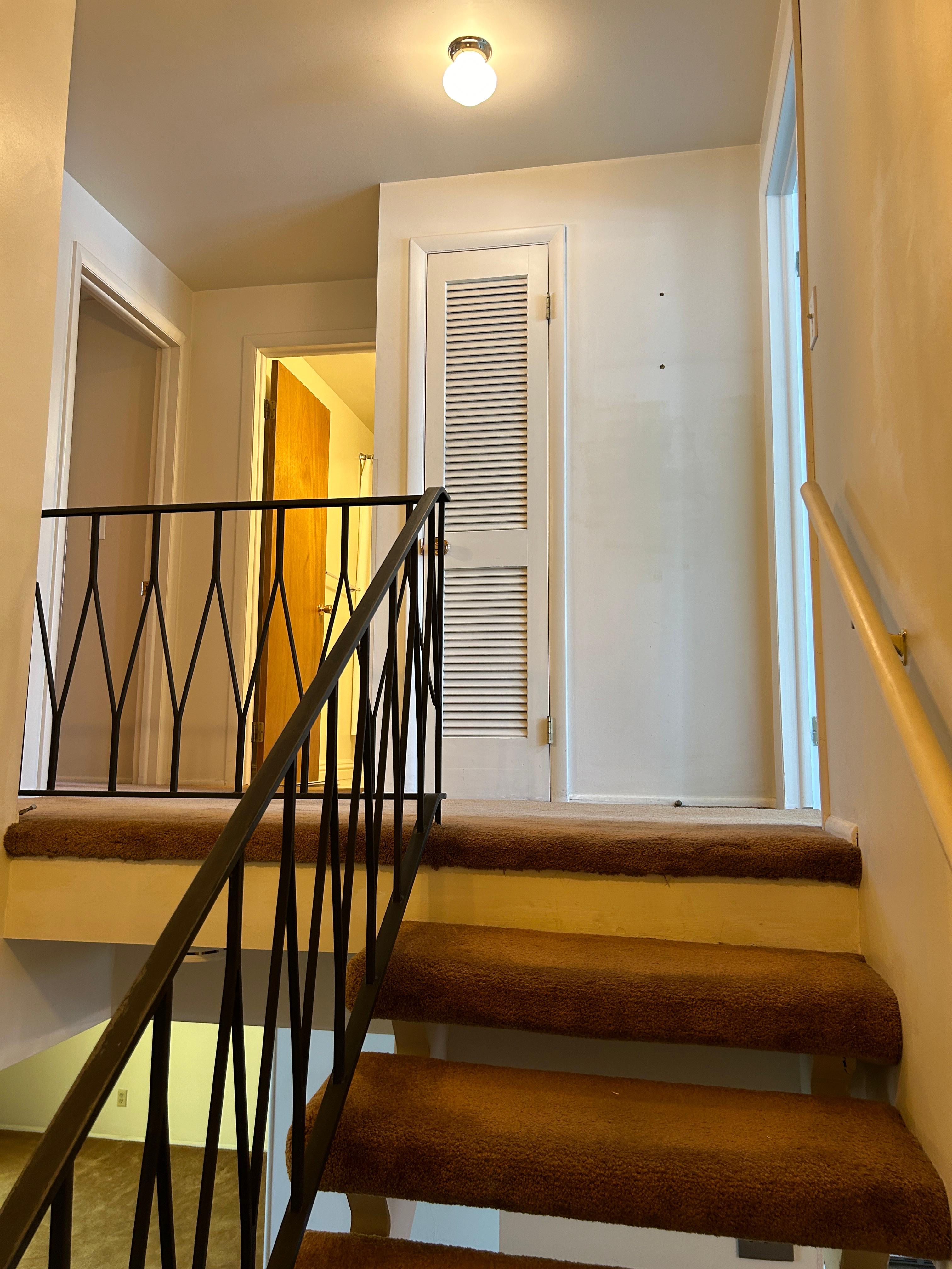 Staircase with brown carpeted treads and risers, featuring a black wrought iron railing and a painted wood handrail