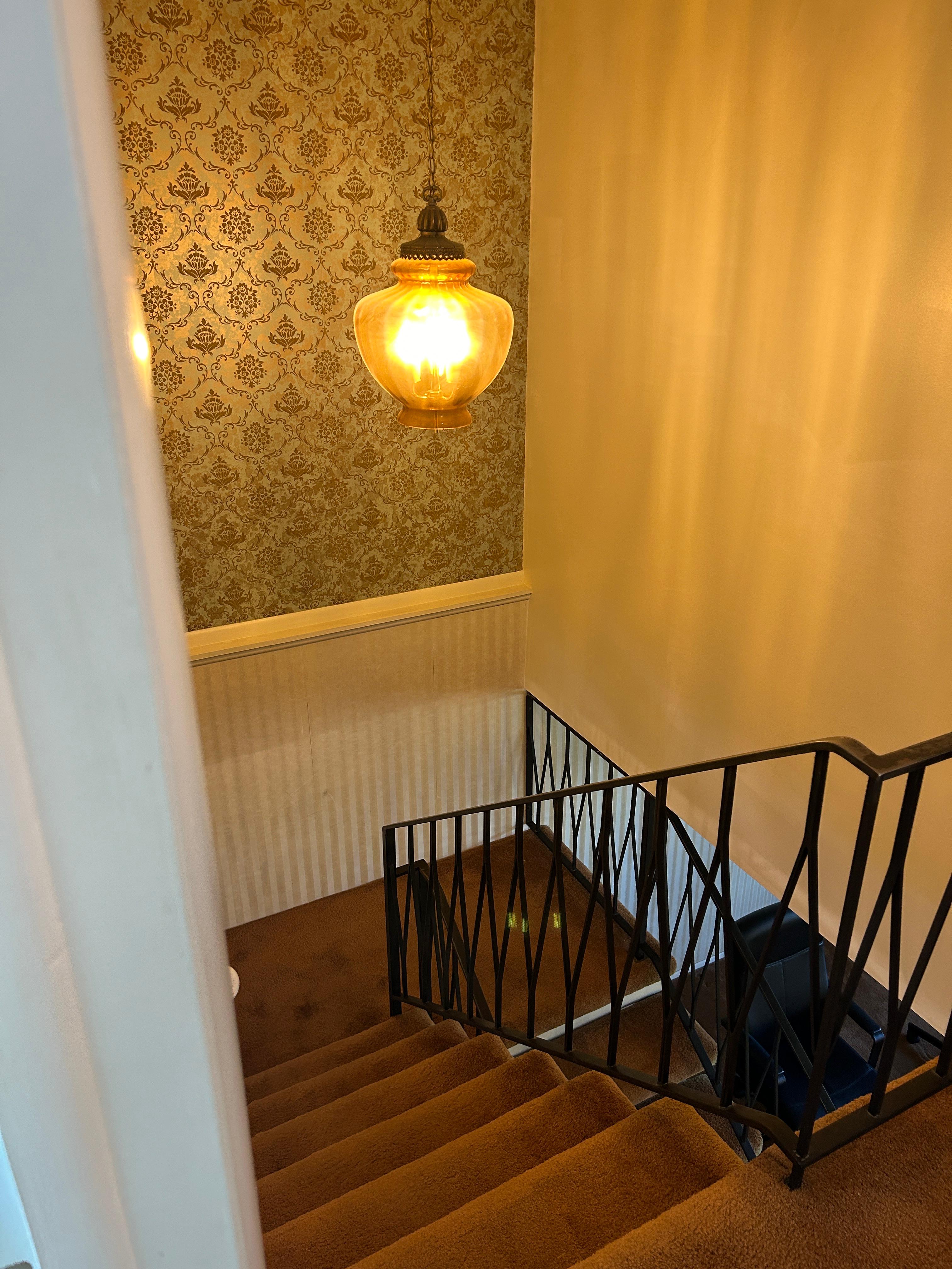 Stairway featuring patterned wallpaper, a decorative pendant light fixture, a black metal railing, and carpeted steps