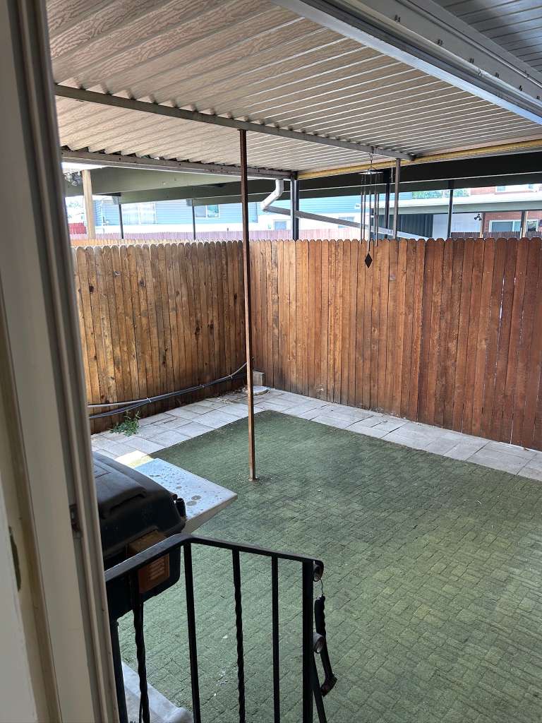 Covered patio area featuring a corrugated metal roof, wood privacy fencing, and a faux grass surface