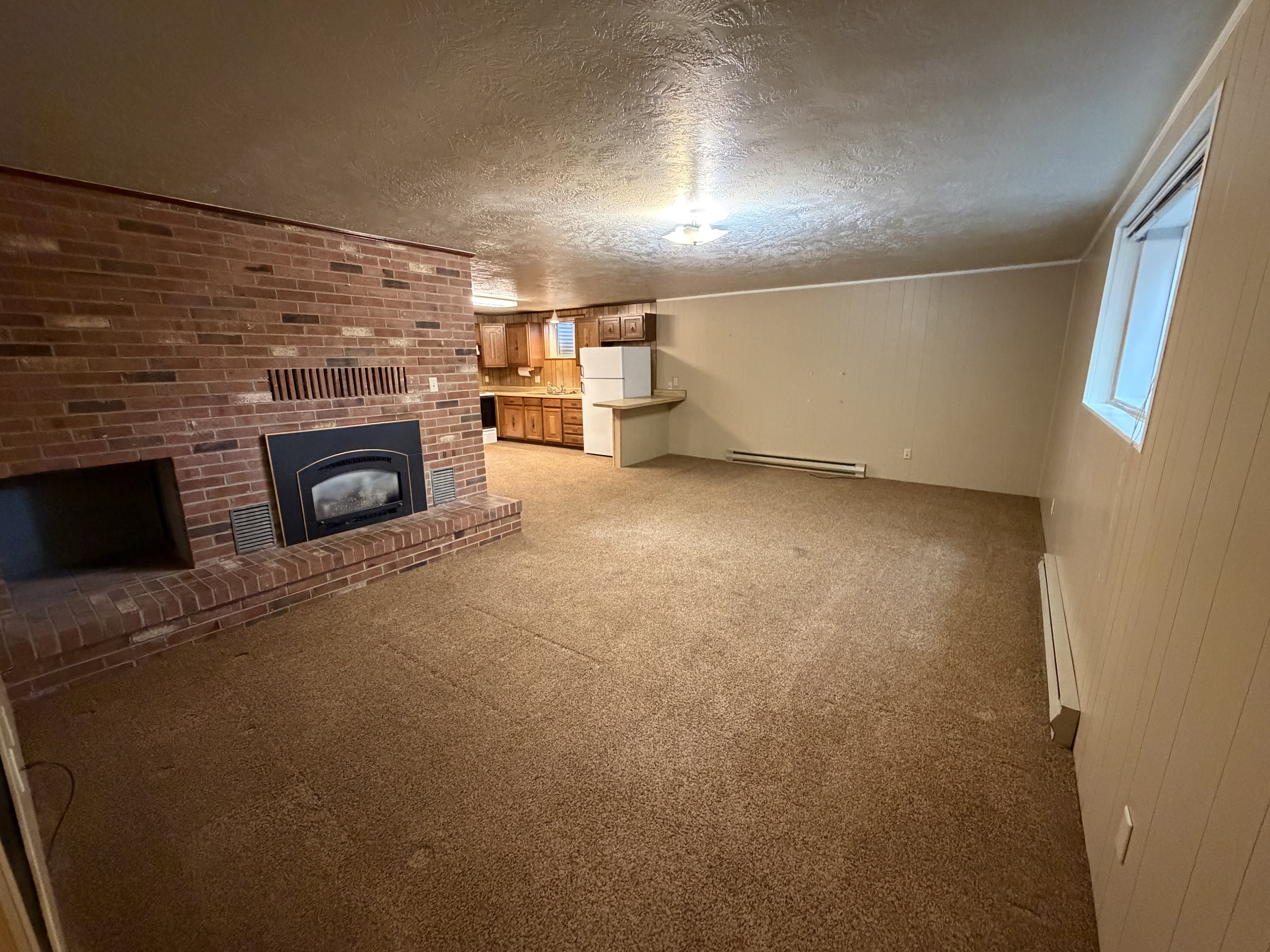Unfurnished living room with light colored carpet, a brick fireplace, a textured ceiling, baseboard heating, and wooden walls