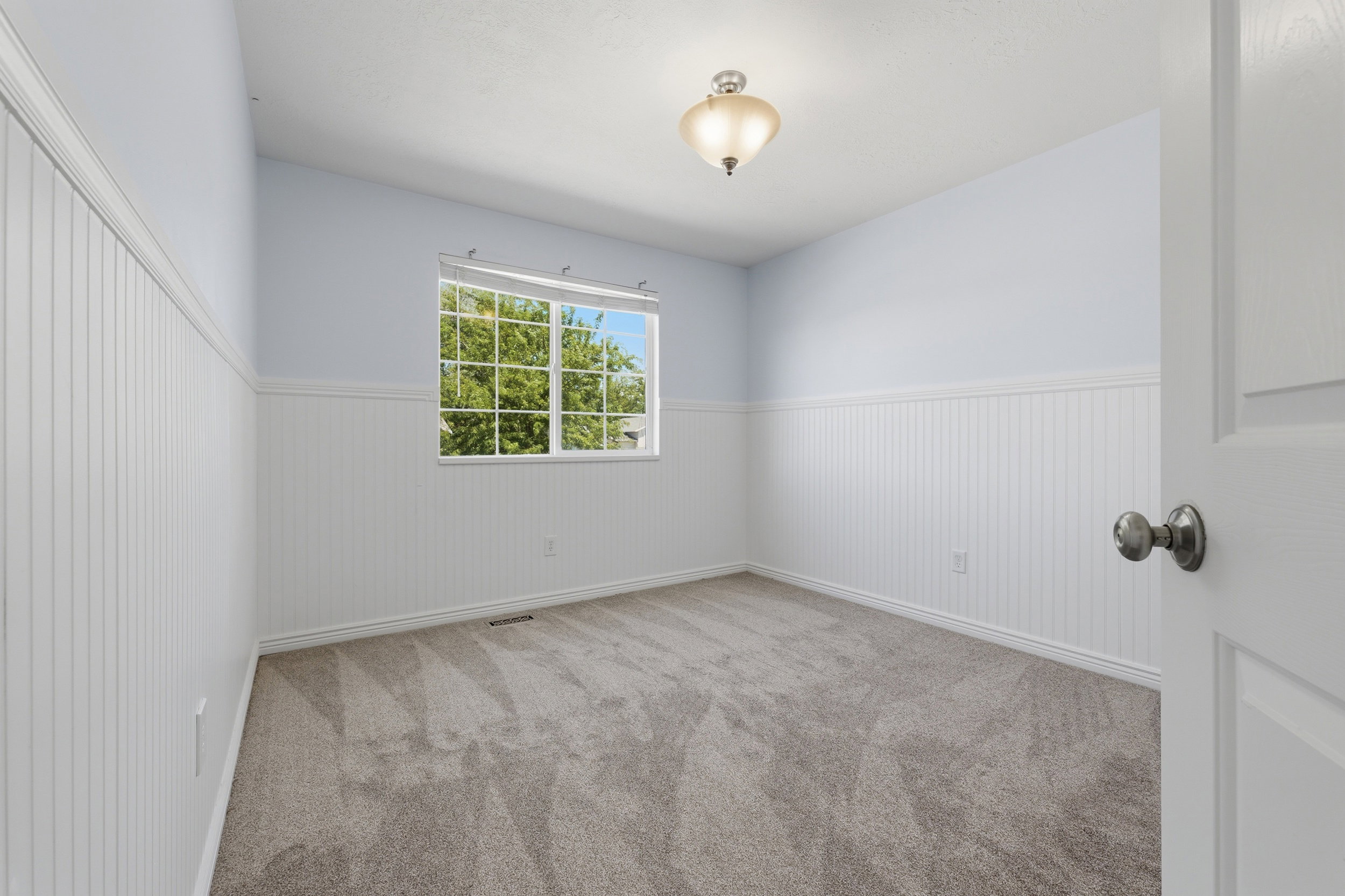 Light-filled room featuring a window with exterior tree views, white beadboard wainscoting, light blue upper walls, and gray carpet flooring