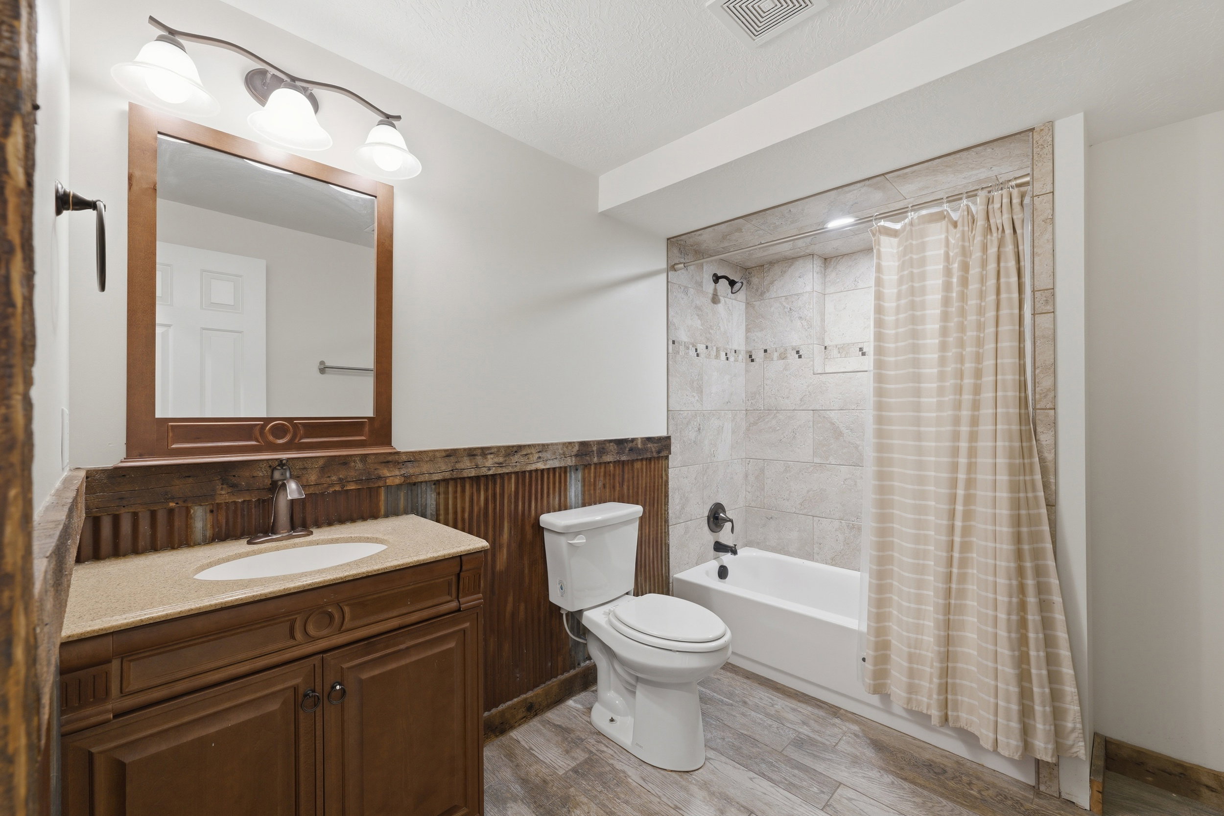 Bathroom featuring a wood-finish vanity with an integrated sink and light-toned countertop, a tiled shower/tub combination with dark fixtures, corrugated metal wainscoting, a framed wall mirror, and wood-look flooring