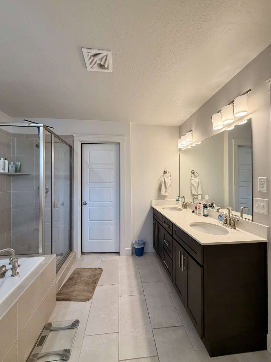 Spacious bathroom featuring a double vanity with dark wood cabinetry and light-colored countertops, large vanity mirror, modern light fixtures, and a shower with glass enclosure