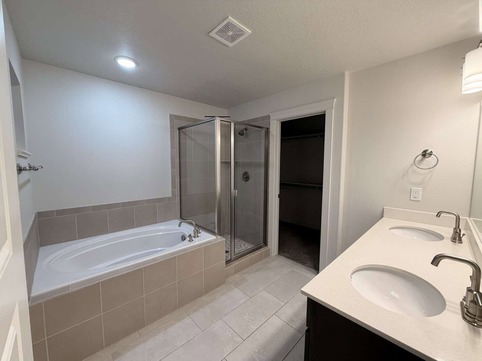 Bathroom featuring a dual-sink vanity with light-toned countertops, a built-in soaking tub with tiled surround, and a glass-enclosed shower