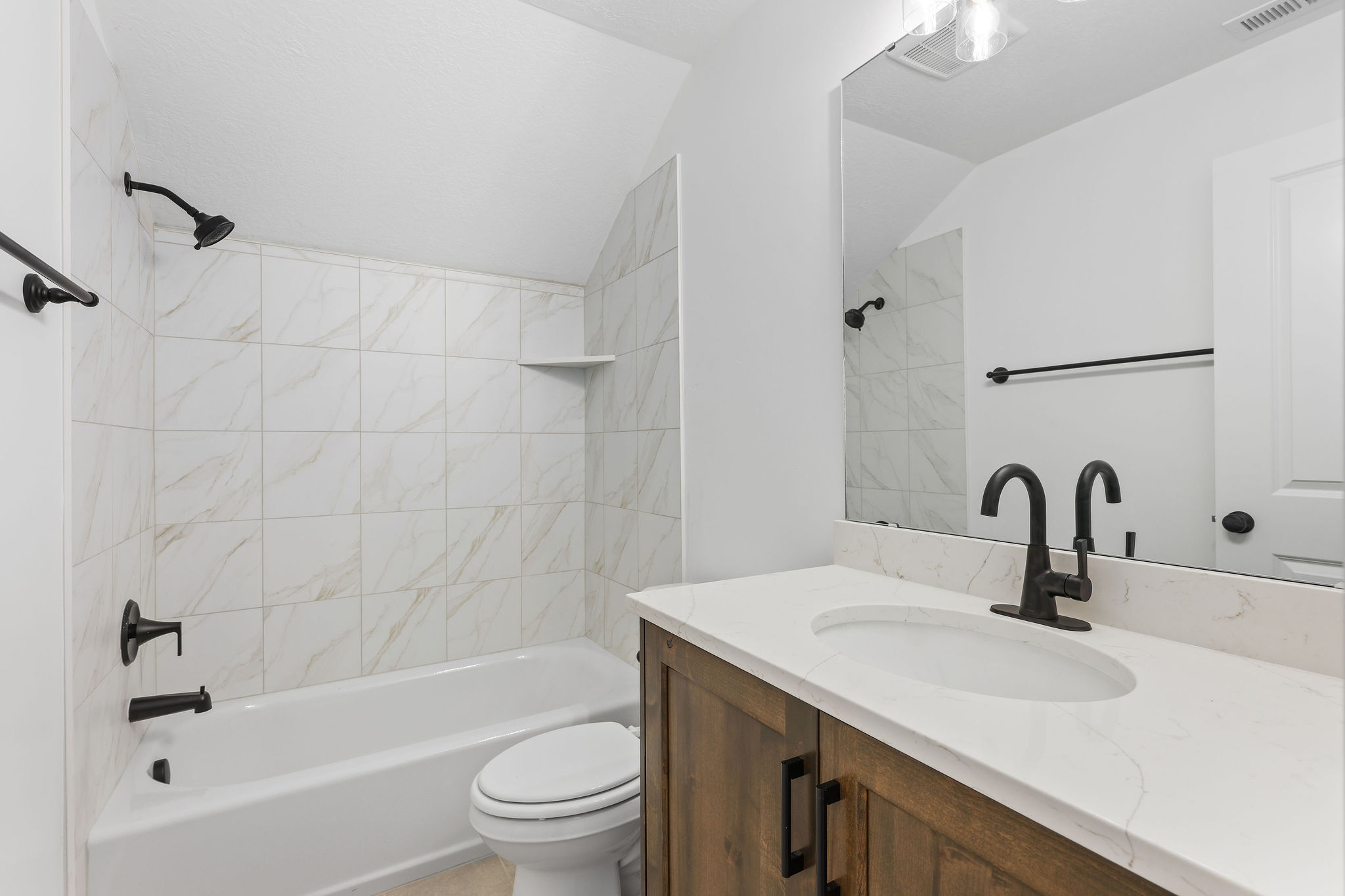 Bathroom featuring a white bathtub with a tiled surround, a wood-finish vanity with a white countertop, a single undermount sink, and matte black fixtures