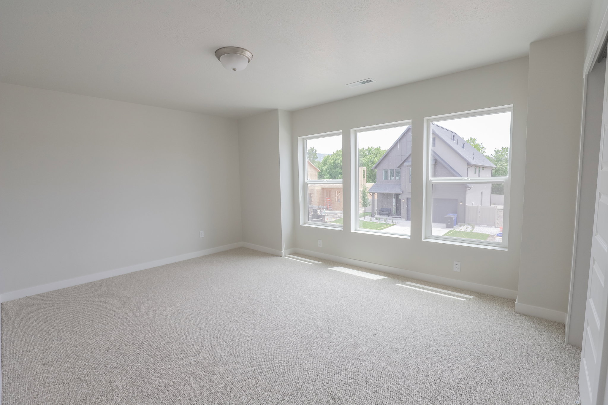 Bright room featuring three large windows, neutral wall paint, light-toned carpeting, and a flush-mount ceiling light fixture