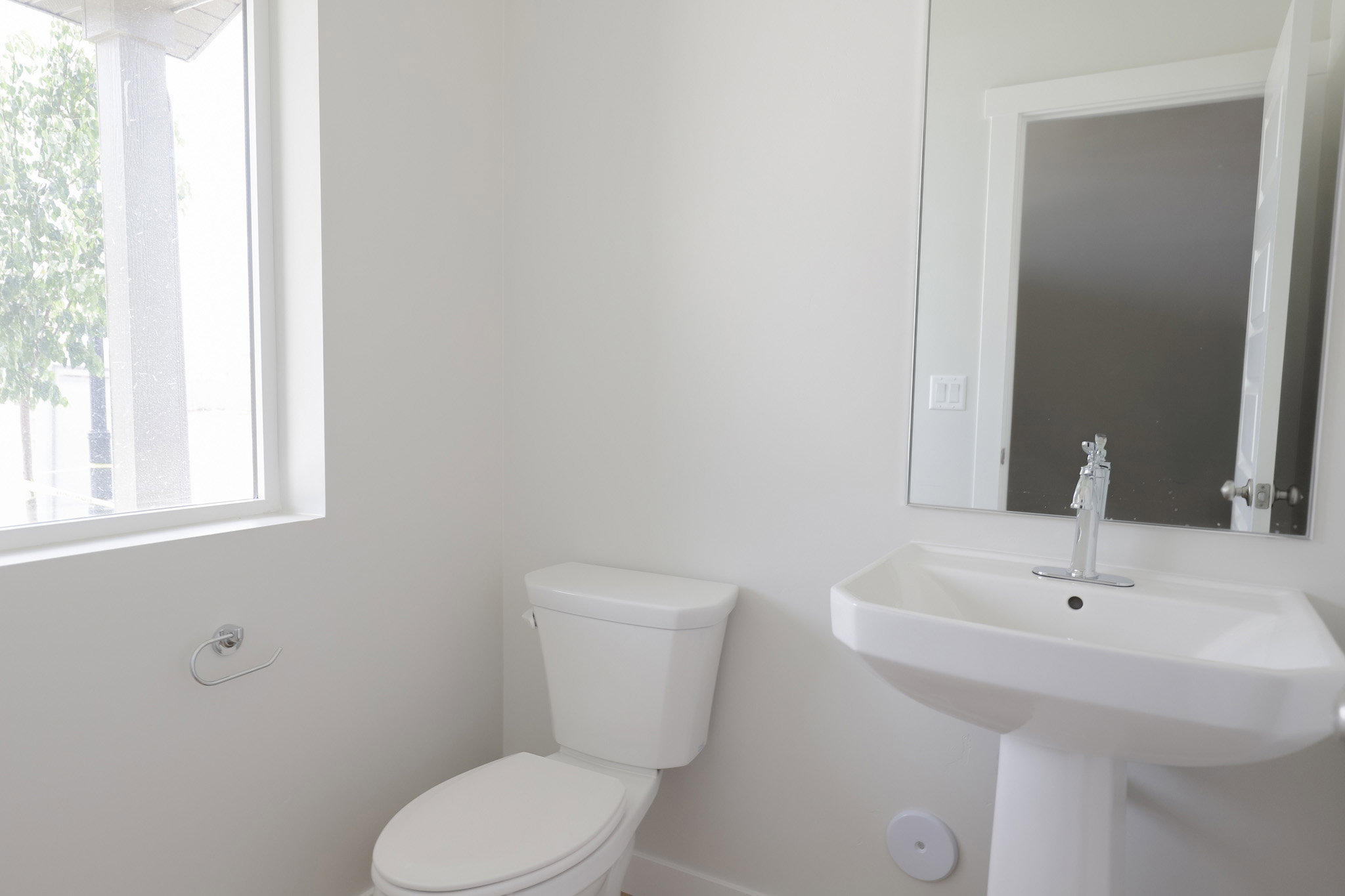 Bathroom featuring a pedestal sink with chrome faucet, toilet, wall-mounted mirror, and a window providing natural light