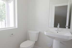 Bathroom featuring a pedestal sink with chrome faucet, toilet, wall-mounted mirror, and a window providing natural light
