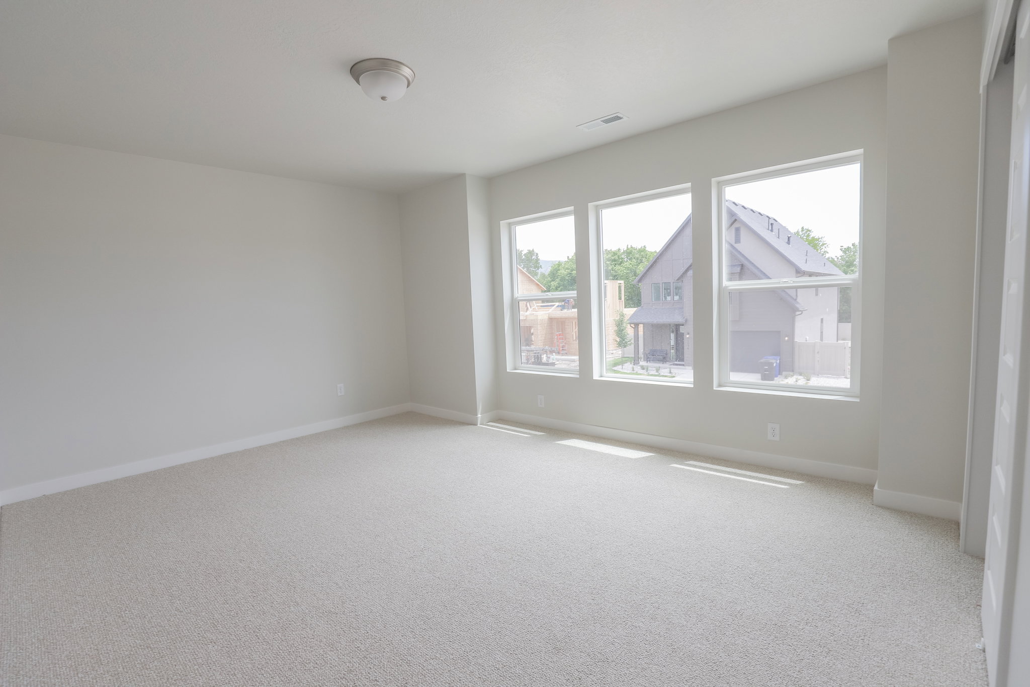 Bright interior space featuring three large windows, light-colored carpeting, white baseboards, and a flush mount ceiling light fixture