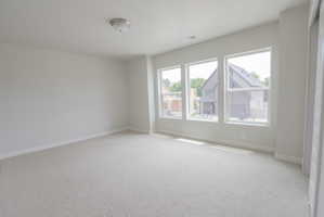 Bright interior space featuring three large windows, light-colored carpeting, white baseboards, and a flush mount ceiling light fixture