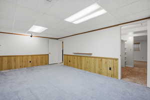 Carpeted empty room featuring wood walls, ornamental molding, a drop ceiling, and a wainscoted wall
