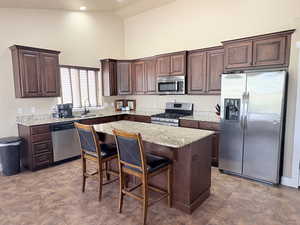 Kitchen featuring stainless steel appliances, dark wood finish cabinetry, a breakfast bar, light stone countertops, and a kitchen island
