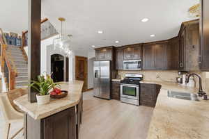 Kitchen with dark wood finish cabinetry, light stone countertops, stainless steel appliances, decorative light fixtures, and light wood-type flooring