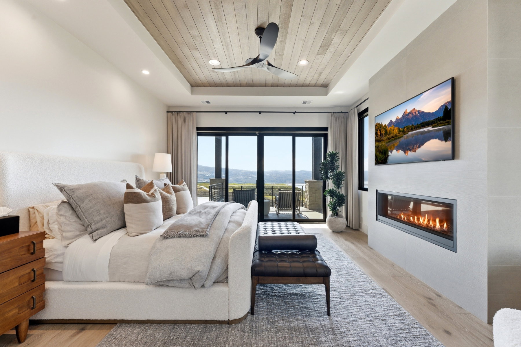 Bedroom featuring access to exterior, a wood tray ceiling, light wood finished floors, a glass covered fireplace, and a ceiling fan