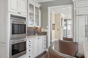 Kitchen featuring double oven, white cabinets, light stone counters, ornamental molding, and glass insert cabinets