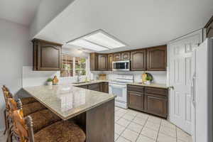 Kitchen featuring dark wood finish cabinets, a breakfast bar, a peninsula, light countertops, and white appliances