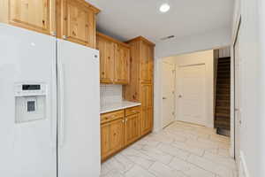Kitchen featuring white fridge with ice dispenser, light countertops, light marble finish flooring, wood finish cabinetry, and backsplash