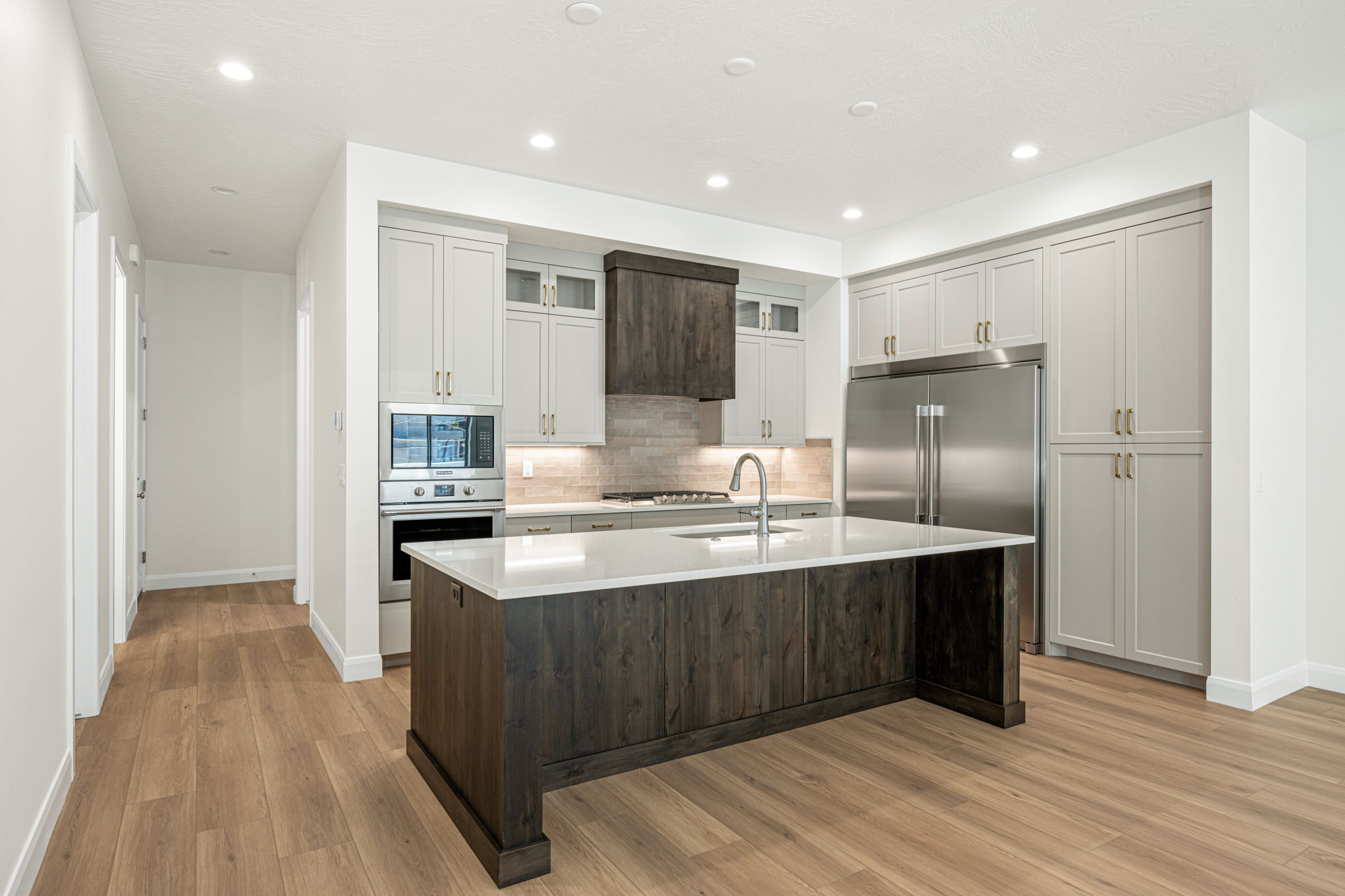 Modern kitchen featuring a large central island with a light-toned countertop, wood-finish flooring, recessed lighting, stainless steel appliances, and two-tone cabinetry with gold-finish hardware