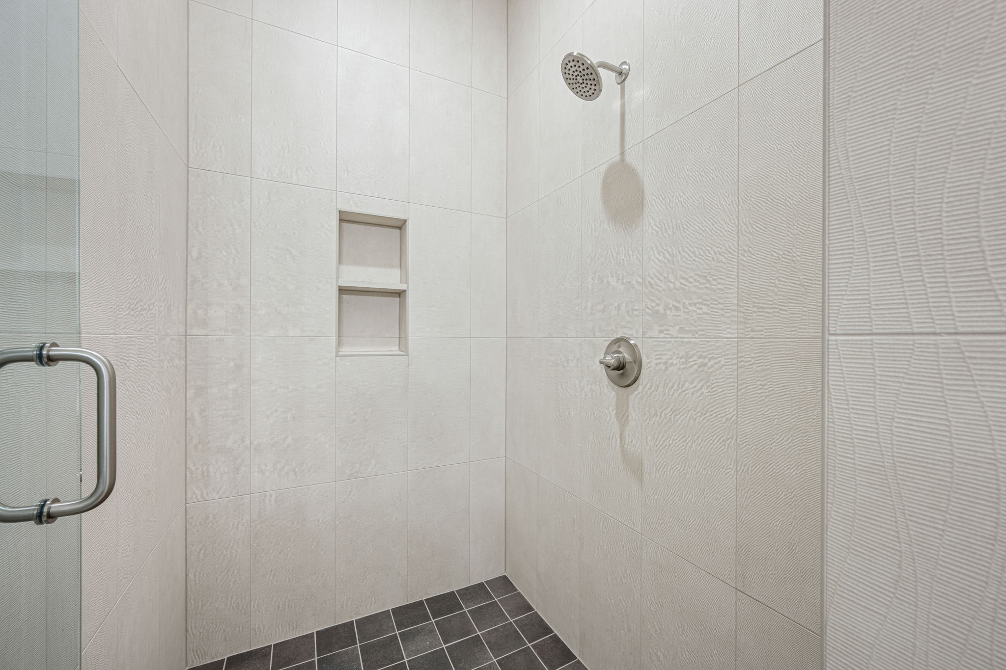 Walk-in shower with light-toned rectangular wall tiles, a built-in shower niche, a brushed nickel shower head and control, and dark square floor tiles