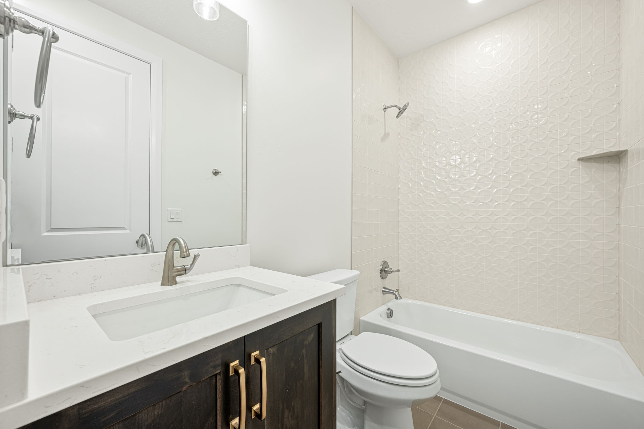 Bathroom featuring a vanity with a white countertop and rectangular undermount sink, dark wood-finish cabinetry with brass-tone hardware, a full-width mirror, and a tub-shower combination with textured white wall tile and an integrated niche