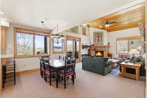 Dining area with light colored carpet, hanging lights, a stone fireplace, ceiling fan, and a vaulted wooden ceiling