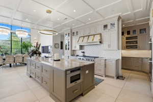 Kitchen with coffered ceiling, light stone counters, two tone color scheme, a center island, and backsplash