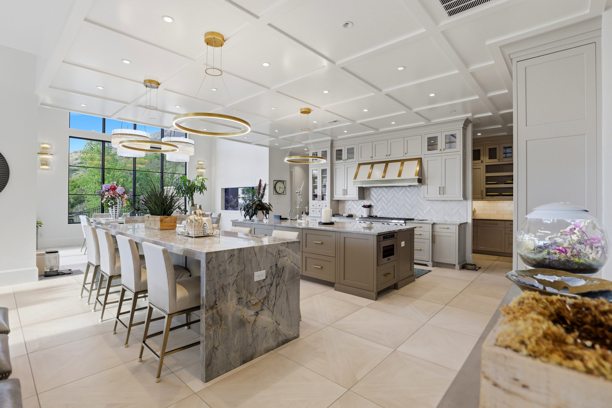 Kitchen with light stone countertops, coffered ceiling, light tile patterned flooring, hanging light fixtures, and glass insert cabinets