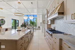 Kitchen with light stone counters, a large island, coffered ceiling, stainless steel gas stovetop, and two tone cabinetry