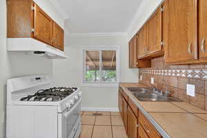Kitchen featuring white gas stove, crown molding, tile counters, wood finish cabinetry, and light tile patterned floors