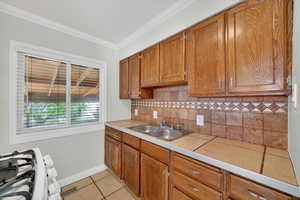 Kitchen with tile counters, wood finish cabinetry, gas range, ornamental molding, and light tile patterned floors