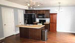 Kitchen with dark wood finish cabinets, black appliances, light countertops, an island with a sink, and dark wood-type flooring