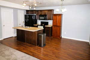 Kitchen with dark wood finish cabinets, black appliances, light countertops, an island with a sink, and dark wood-type flooring