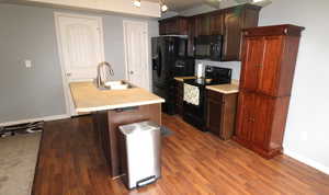 Kitchen with dark wood finish cabinets, black appliances, light countertops, an island with a sink, and dark wood-type flooring