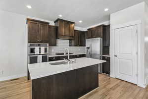 Kitchen featuring dark wood finish cabinets, light stone counters, a kitchen island with sink, light wood finished floors, and stainless steel appliances