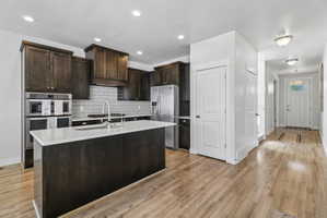 Kitchen featuring dark wood finish cabinets, stainless steel appliances, light stone counters, a center island with sink, and light wood finished floors