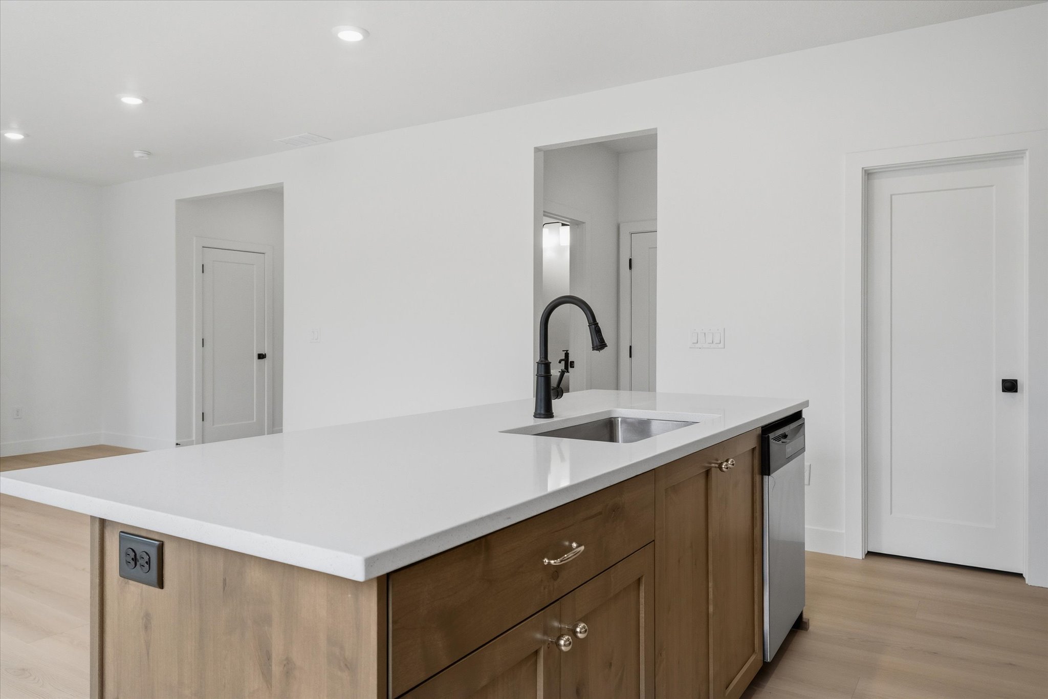 Kitchen island featuring a white countertop, undermount sink with a matte black gooseneck faucet, light wood-finish cabinetry, and an integrated stainless steel dishwasher
