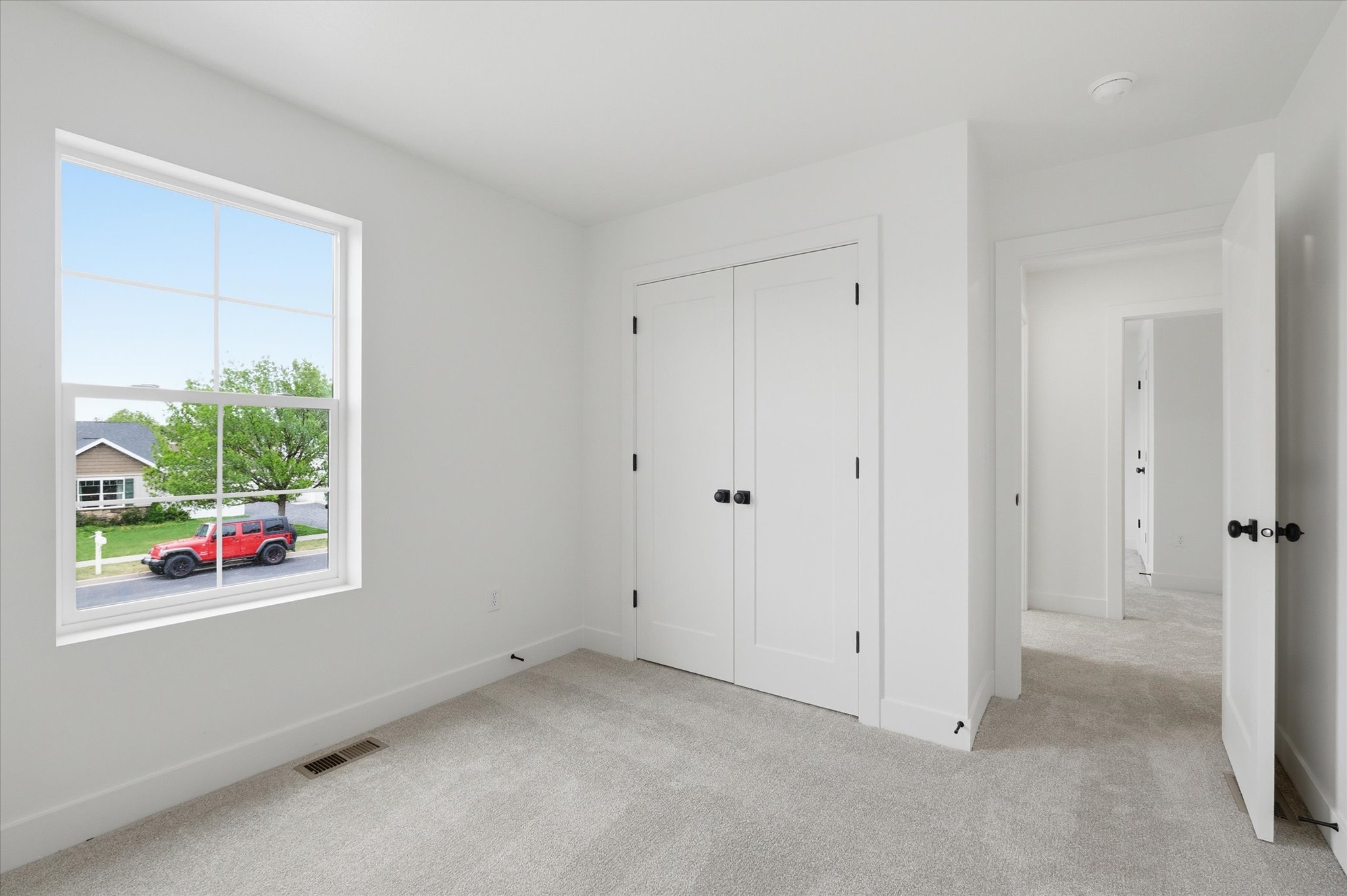 Bright interior room featuring a large window, light-toned carpeting, white walls, and a double-door closet with black hardware