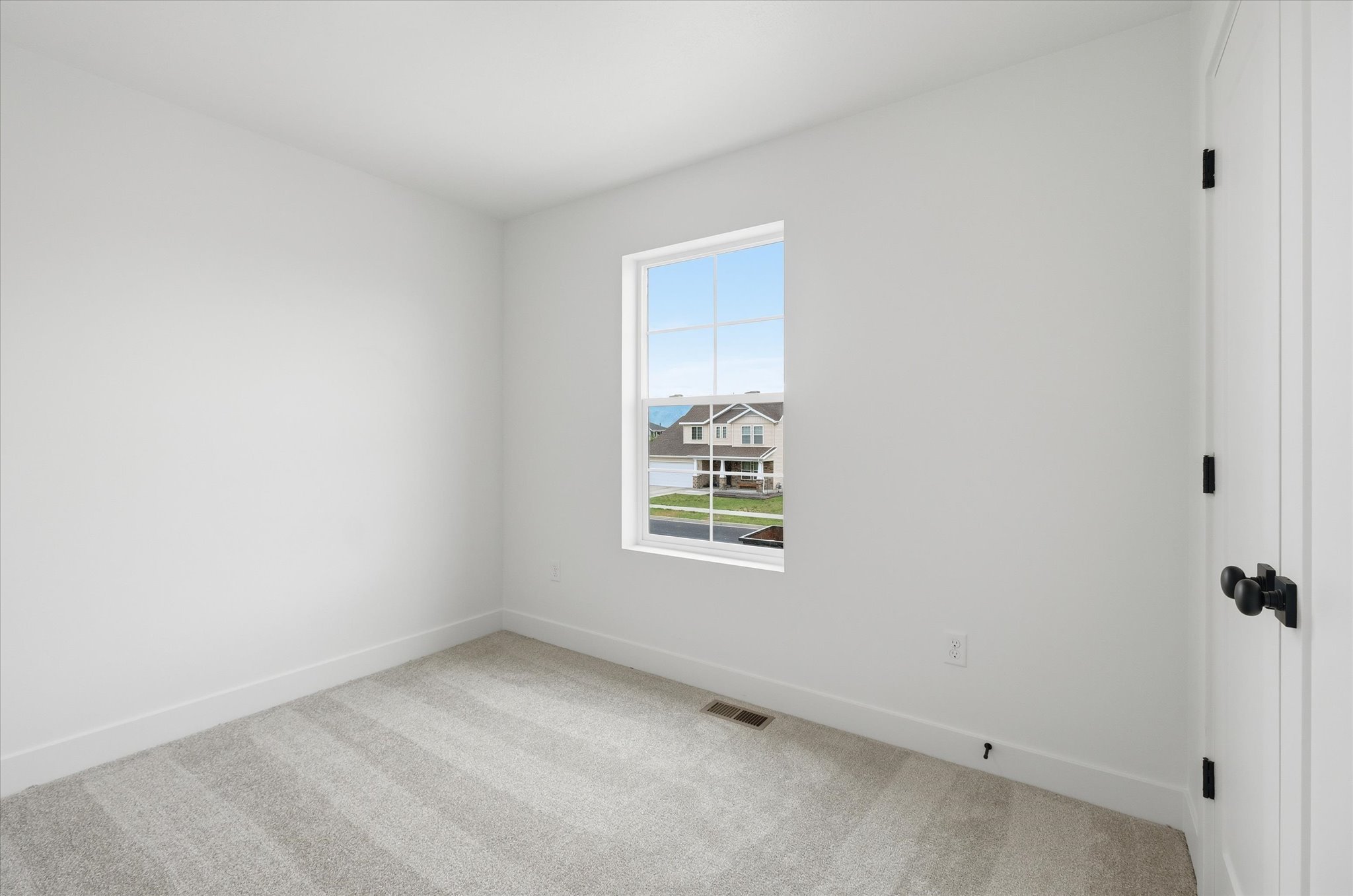 Bright interior space featuring a window with grid panes, light-colored carpeting, white baseboards, and a white interior door with matte black hardware